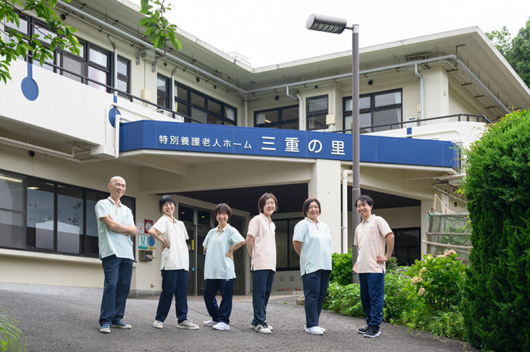 介護施設の玄関に職員6名が並んで立っている。建物外観と入口が見える。