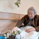 居室でくつろぐ高齢者女性。窓際で花を飾り居心地よく整備された空間。